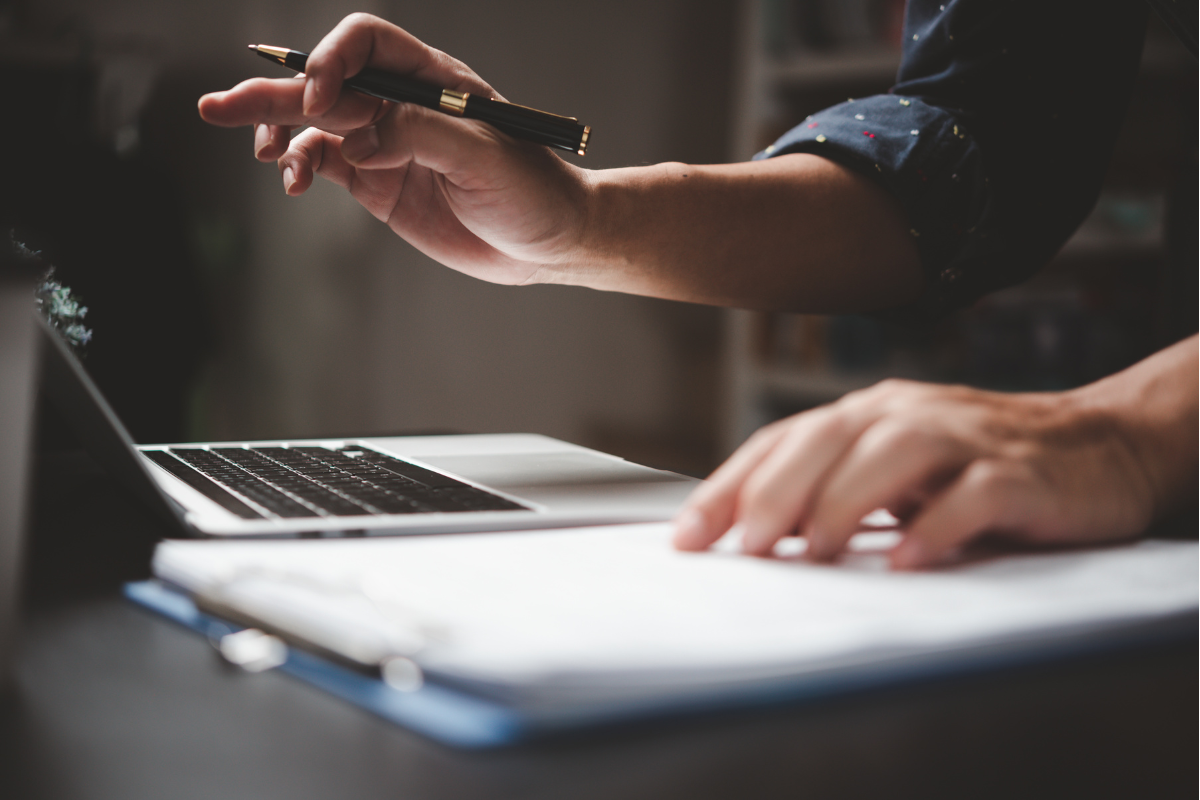 an open laptop and paperwork on a desk with human hands holding a pen